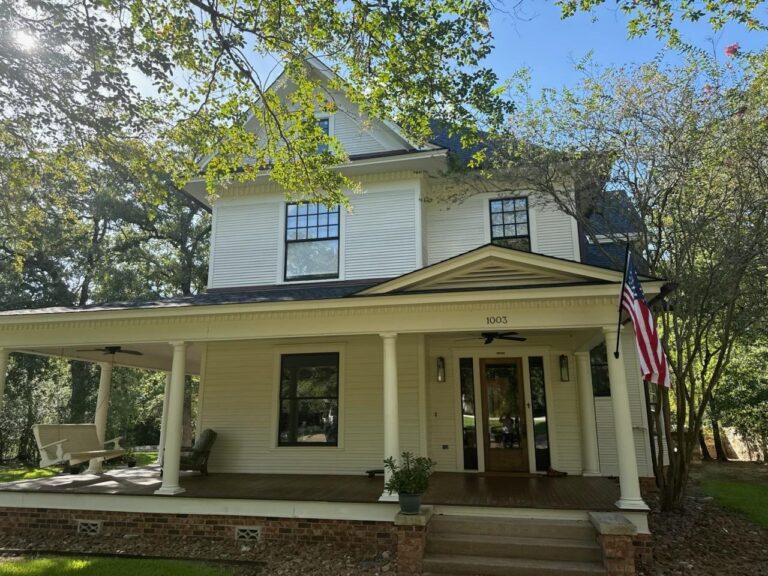 Repainted two-storey white farmhouse with a wraparound porch and dark window trim in the Brazos Valley, Texas